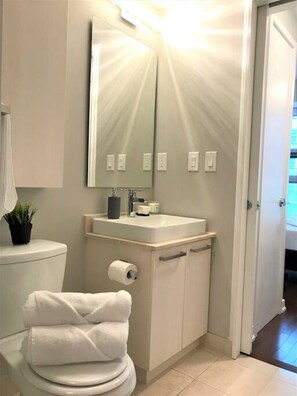 Ein Badezimmer mit Toilette, Waschbecken und Spiegel. Auf dem Toilettendeckel liegt ein Handtuch und auf dem Waschbecken eine Pflanze.
