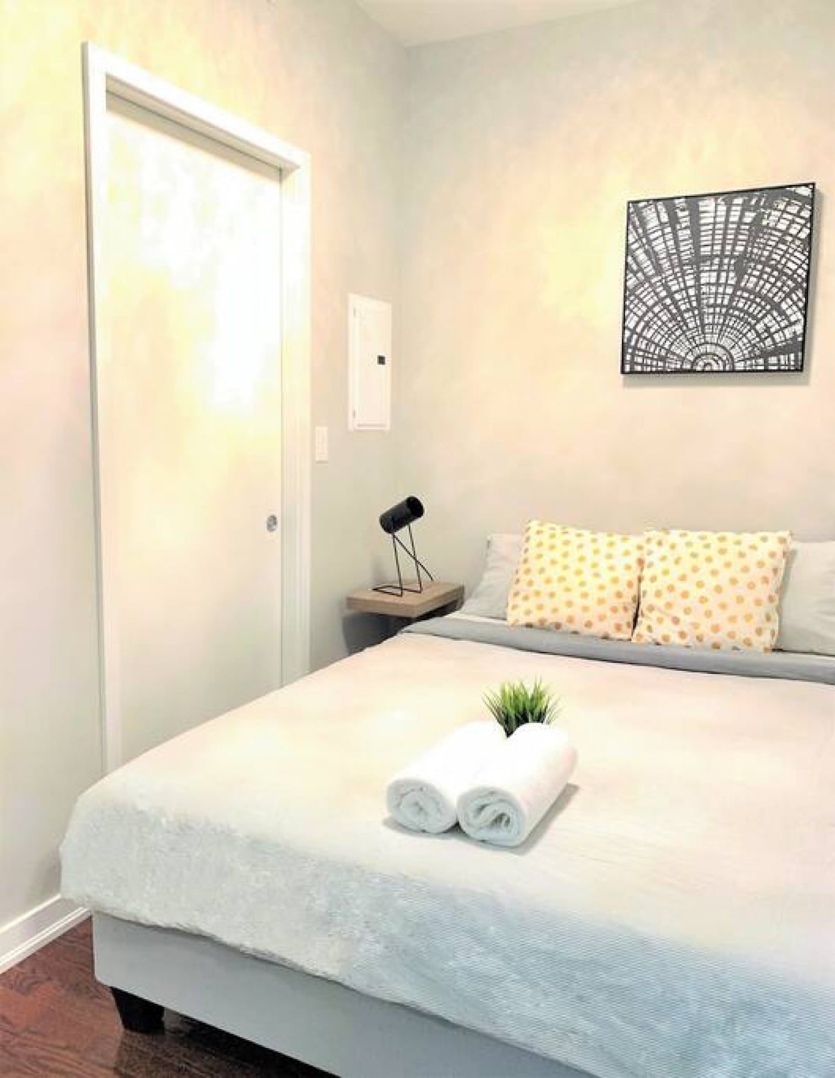 A neatly made bed with a rolled towel and a small plant, a bedside lamp, and a wall art piece.