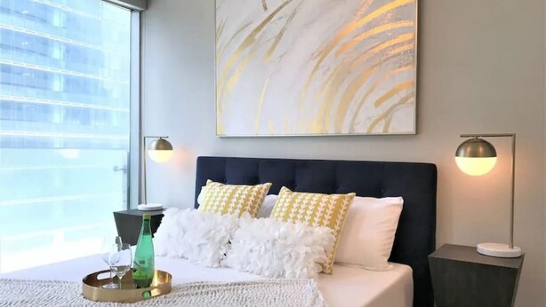 A modern bedroom with a large bed, decorative pillows, a wall art piece, and a view of a building through a window. - Downtown Condo with Juliette Balcony and Parking (Toronto)