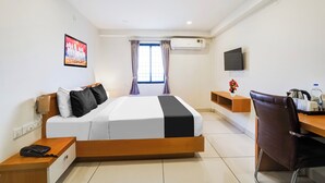 Quarto de hotel com cama, escrivaninha, cadeira, televisão, ar condicionado e janela com cortinas.