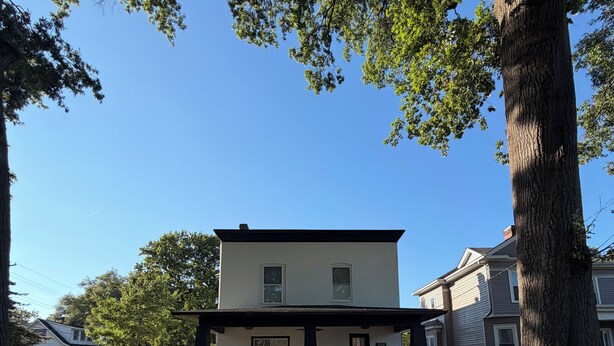 Uma casa de dois andares com varanda da frente, entrada por uma rampa e uma grande árvore ao primeiro plano.