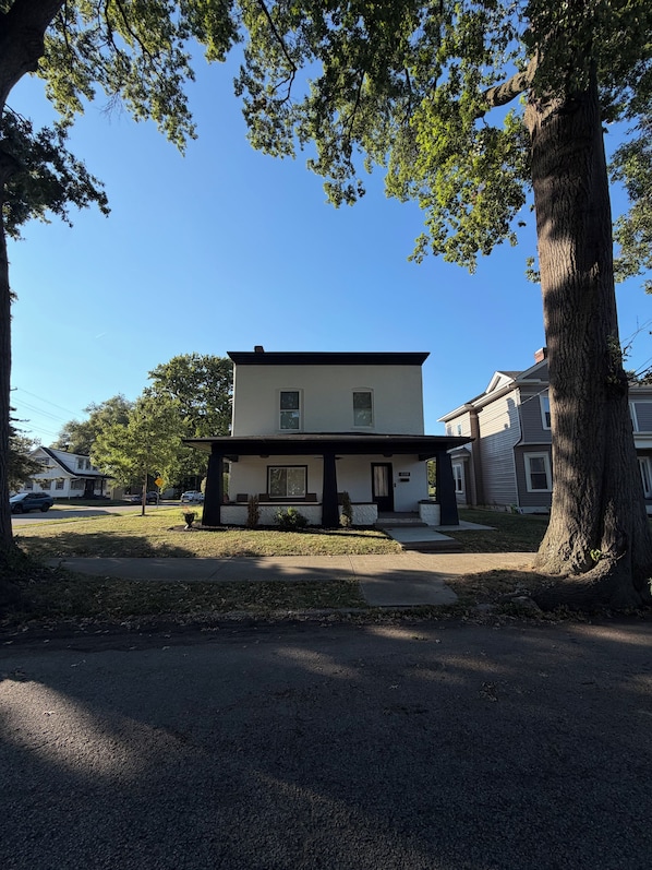 A two-story house with a front porch, a driveway, and a large tree in the foreground.