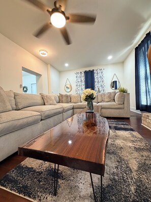 A living room with a sectional sofa, a wooden coffee table, a ceiling fan, and a large window with curtains.