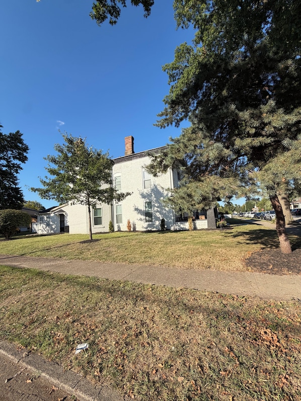 A white building with a brick chimney, surrounded by trees and a grassy lawn.