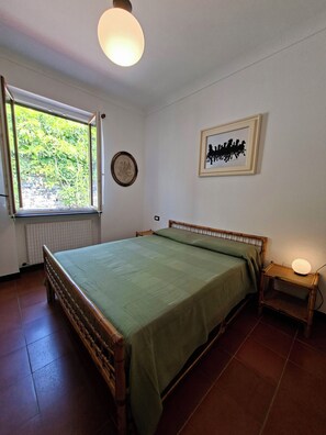 A bedroom with a bed, a bedside table with a lamp, a window with a view of greenery, and a framed picture on the wall. - Oasi Nel Verde Just a few dozen meters from the sea and close to downtown (Rapallo)