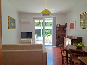 A living room with a sofa, a television on a stand, a dining table with chairs, a bookshelf, and a door leading to a balcony. - Oasi Nel Verde Just a few dozen meters from the sea and close to downtown (Rapallo)