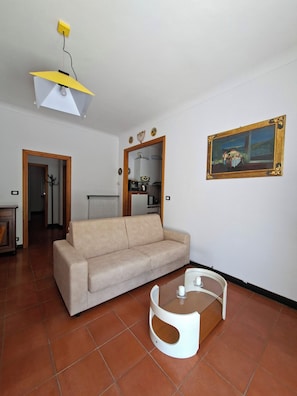 A living room with a sofa, a coffee table, a painting on the wall, and a kitchen area visible through an open doorway. - Oasi Nel Verde Just a few dozen meters from the sea and close to downtown (Rapallo)