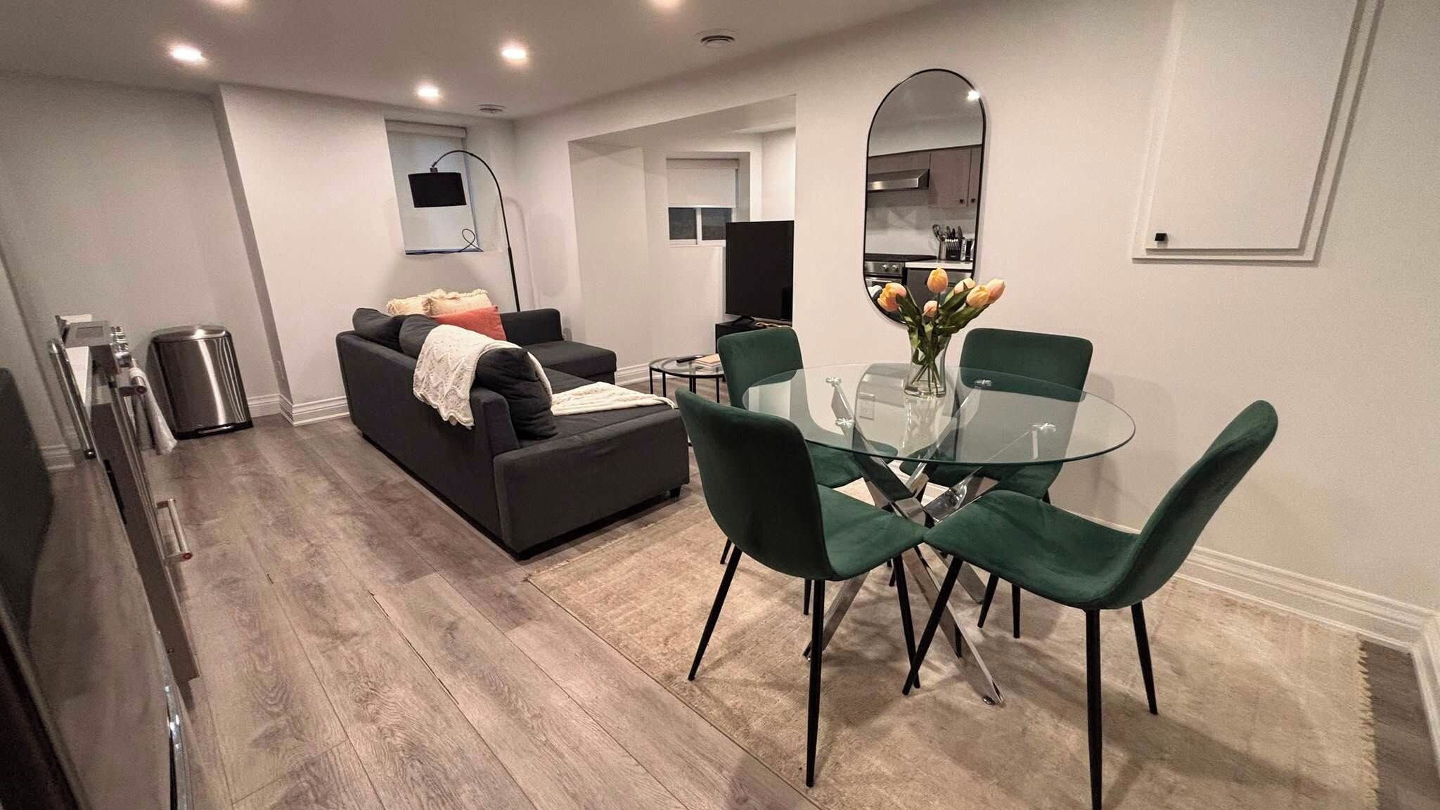 A modern living room with a sofa, a glass dining table with chairs, a kitchen area with a microwave, and a TV mounted on the wall.