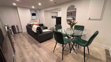 A modern living room with a sofa, a glass dining table with chairs, a kitchen area with a microwave, and a TV mounted on the wall.