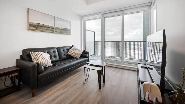 Uma sala de estar moderna com um sofá de couro preto, uma mesa redonda pequena e uma grande janela com vista para a paisagem urbana.