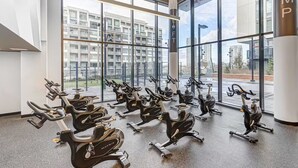 Uma academia com várias bicicletas ergométricas dispostas em fileiras, grandes janelas e vista para a cidade.