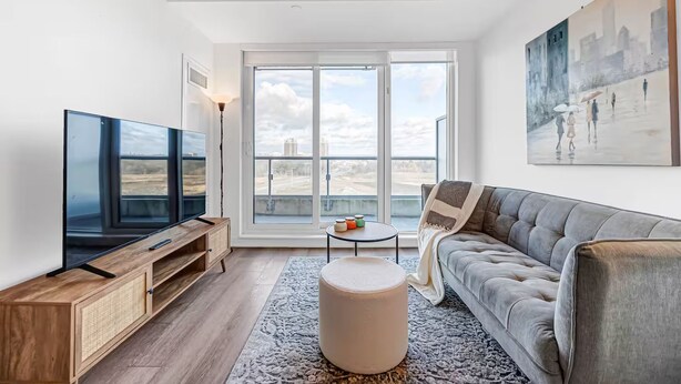 Uma sala de estar moderna com televisão de tela plana, um sofá cinza, uma mesa de centro e uma grande janela com vista para a paisagem urbana.