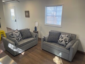 Sala de estar com dois sofás cinzas, almofadas estampadas, uma mesinha com abajur, uma janela com persianas e uma porta.
