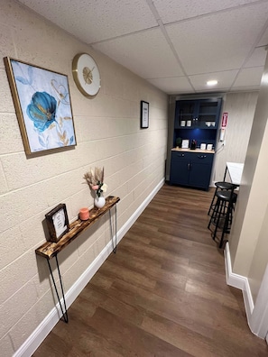 A hallway with a wooden floor, a console table with a vase and a framed picture, a clock on the wall, and a blue cabinet with a fire extinguisher.