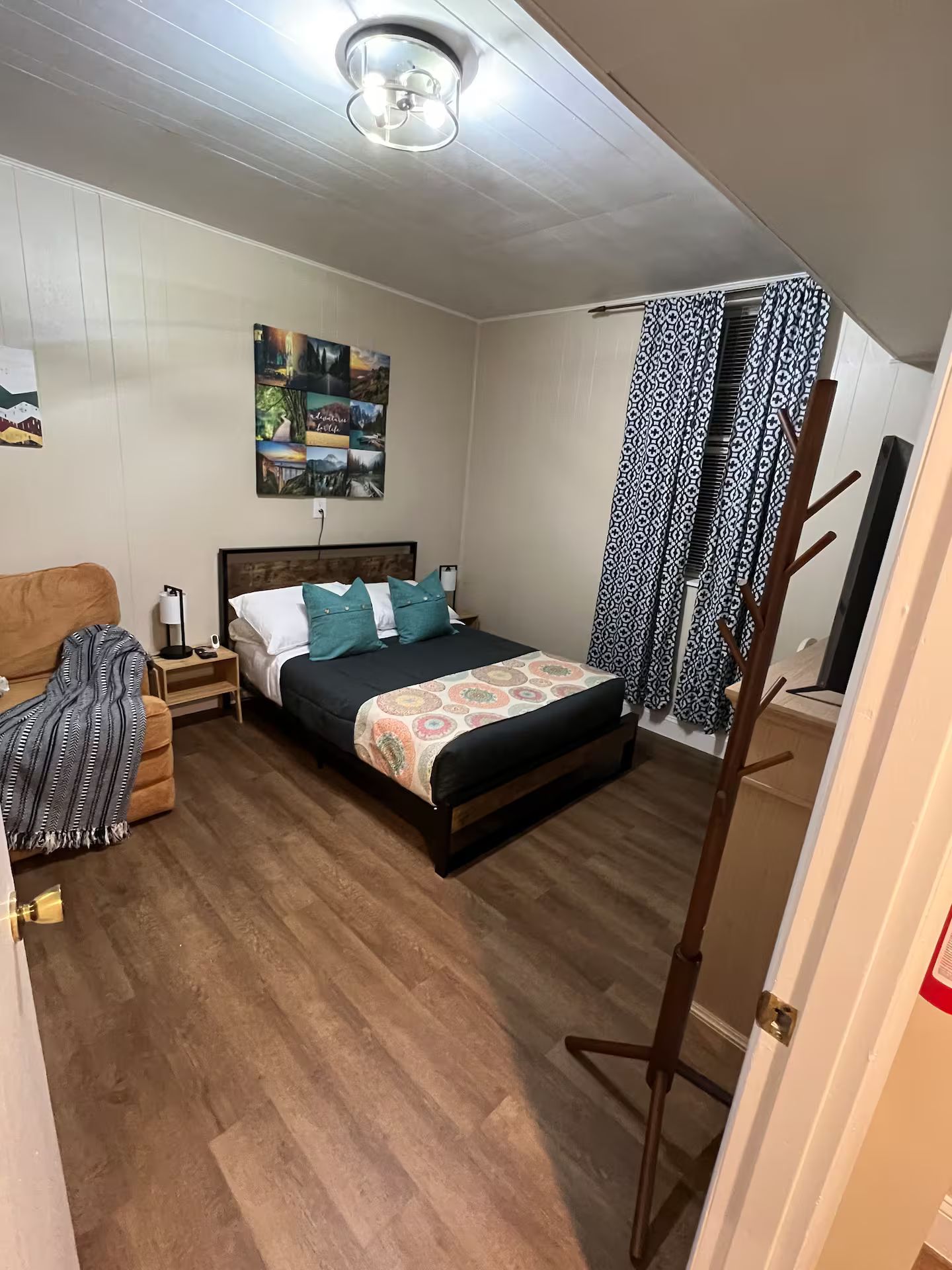 Ein Schlafzimmer mit einem Bett, einem Nachttisch, einer Lampe, einem Wandbild und einem gemusterten Vorhang.