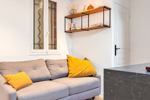 Ein modernes Wohnzimmer mit einem grauen Sofa, gelben Kissen, einem Holzregal mit Büchern und einem Fenster mit Jalousien.