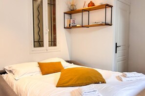 Una cama bien hecha con almohadas blancas y mostaza, un estante con libros y un florero, y una puerta con picaporte.