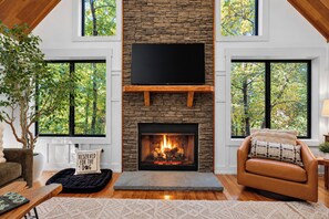 A modern living room with a stone fireplace, a flat-screen TV mounted above it, and a large window on one side. - Wintergreen Luxury! Sleeps 18 with Hot Tub, Sauna, Arcades (Wintergreen Resort)