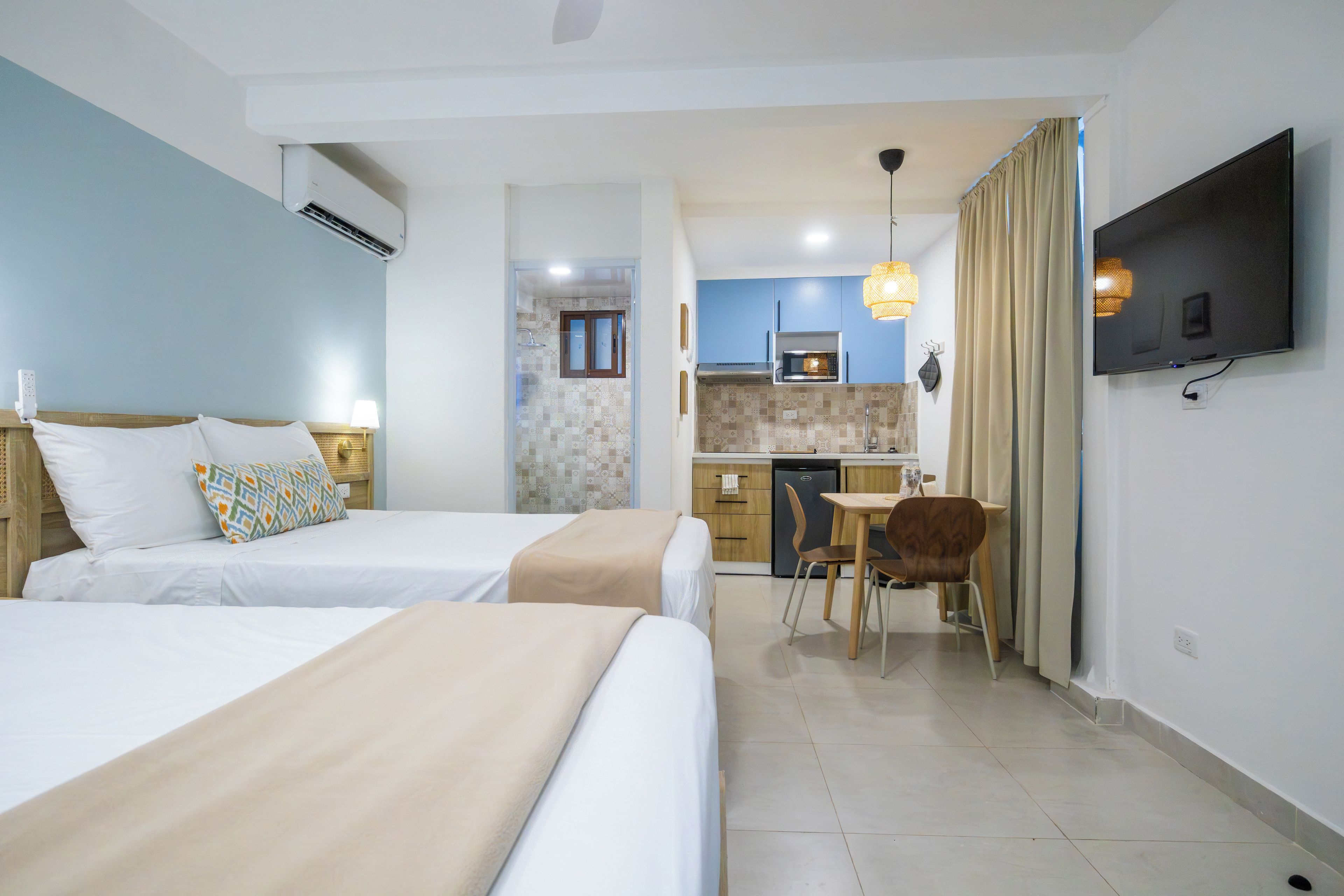 Habitación de hotel con dos camas, kitchenette, zona de comedor y televisor montado en la pared.