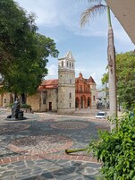 Una plaza con un edificio histórico que cuenta con una torre de campanas y arcos, rodeada de árboles y una estatua.