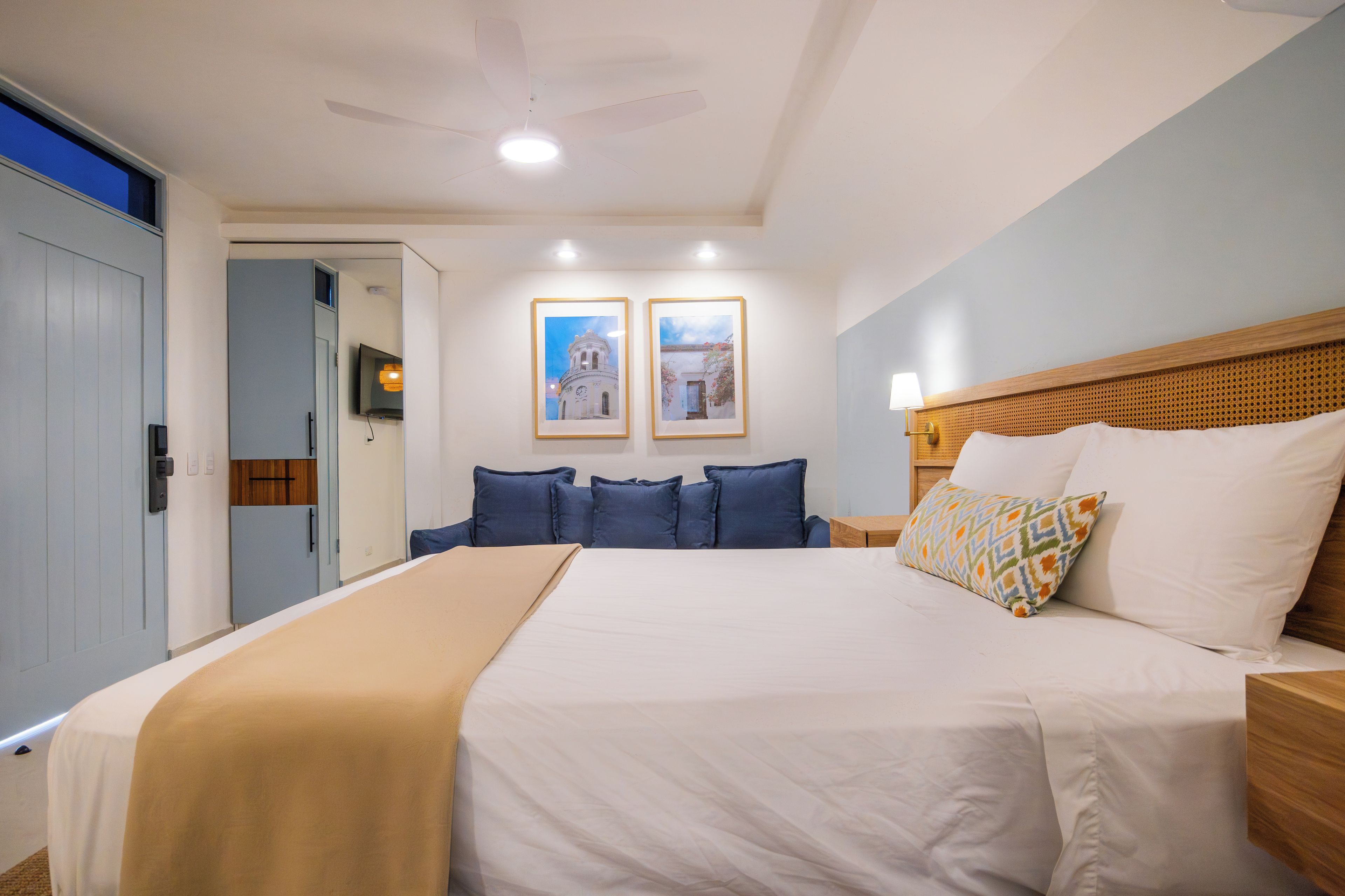 Una habitación de hotel con una cama, un ventilador de techo, un televisor de pantalla plana y dos cuadros en la pared.