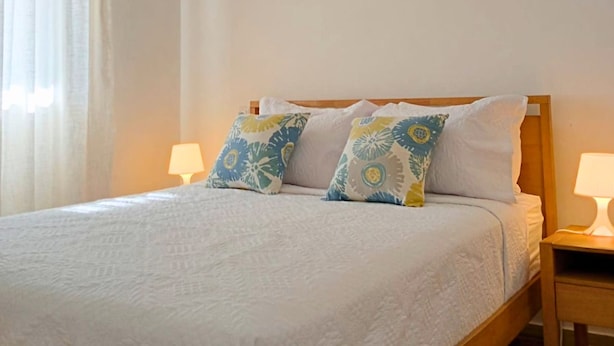 Cama com estrutura de madeira, roupa de cama branca e almofadas estampadas. Duas luminárias de cabeceira sobre mesas de apoio de madeira.