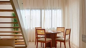 Uma sala de estar moderna com área de jantar, móveis de madeira, escada e grandes janelas com cortinas leves.
