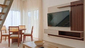 Uma sala de estar moderna com televisão de tela plana fixada na parede, uma mesa de jantar com cadeiras e uma escada visível ao fundo.