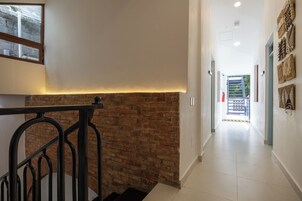 Un interior moderno con pared de ladrillo decorativa, una escalera con barandillas de metal y un pasillo que conduce a una puerta con vistas a zonas verdes.
