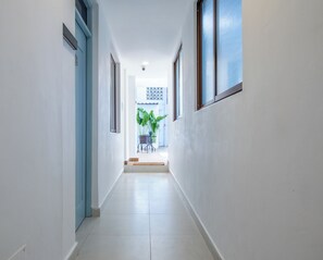 Un pasillo con una puerta a la izquierda, una ventana a la derecha y vista de una habitación con planta y muebles al fondo.