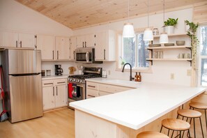 A modern kitchen with white cabinetry, stainless steel appliances, a large island with stools, and a window with a view of trees. - Tranquil Cabin Escape with Private Sauna & Kayak Fun in Mission Township, Minnesota (Mission Township)