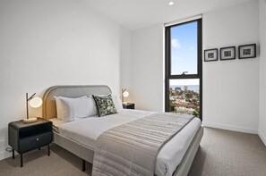 A bedroom with a large bed, two bedside tables, a nightstand, a window with a city view, and three framed pictures on the wall. - Light-filled 3-bed Brand New Apartment (Melbourne)