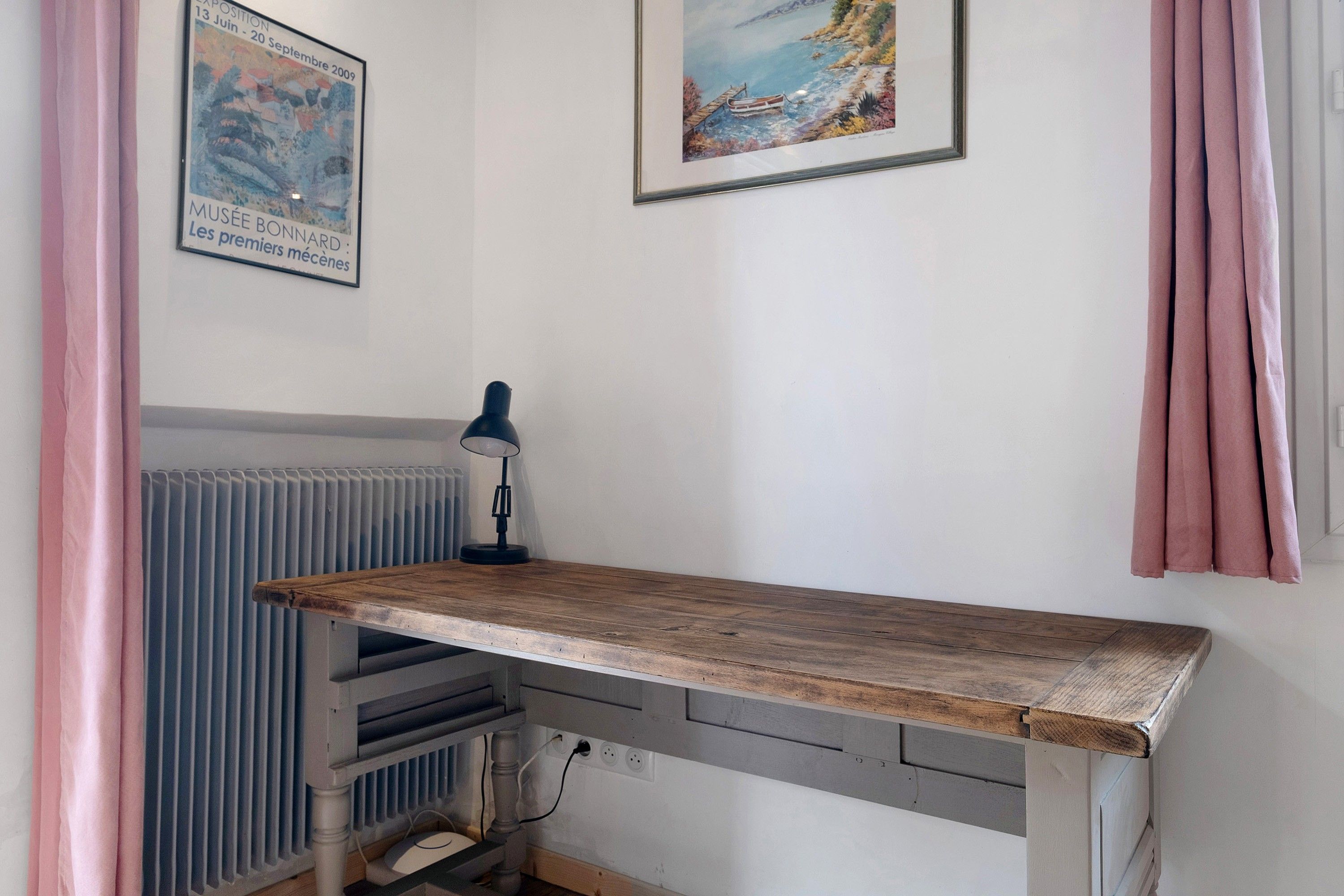 A room with a wooden desk, a radiator, a lamp, and two framed pictures on the wall.