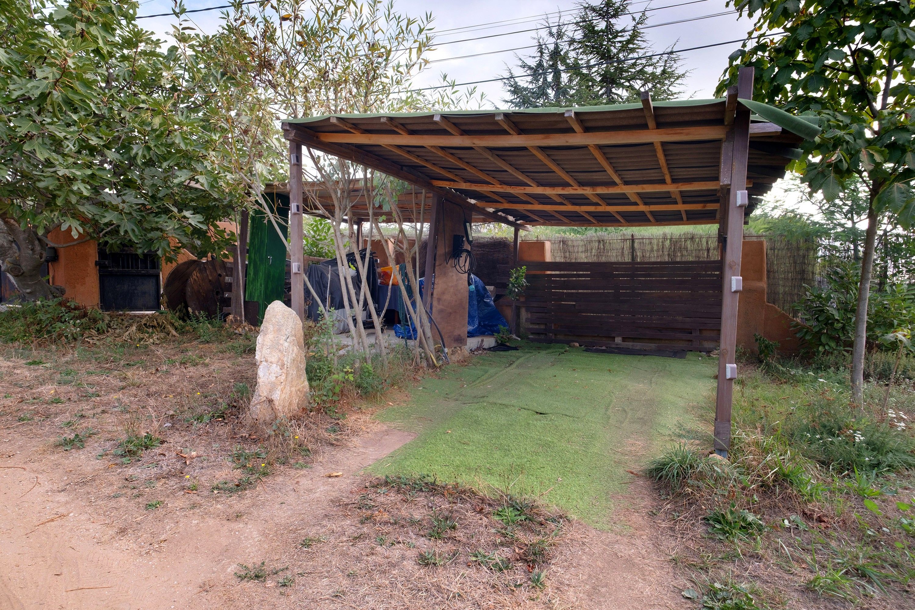 A covered outdoor area with a dirt path leading to a green lawn, surrounded by trees and a wooden fence.
