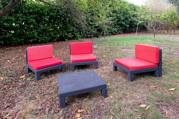 A set of three outdoor wicker chairs with red cushions and a matching ottoman, arranged on a leaf-covered ground.