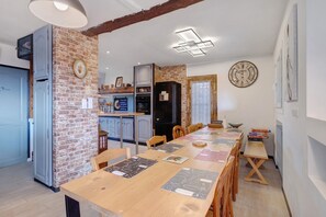 A dining area with a long wooden table, kitchen area with a refrigerator, and a clock on the wall.