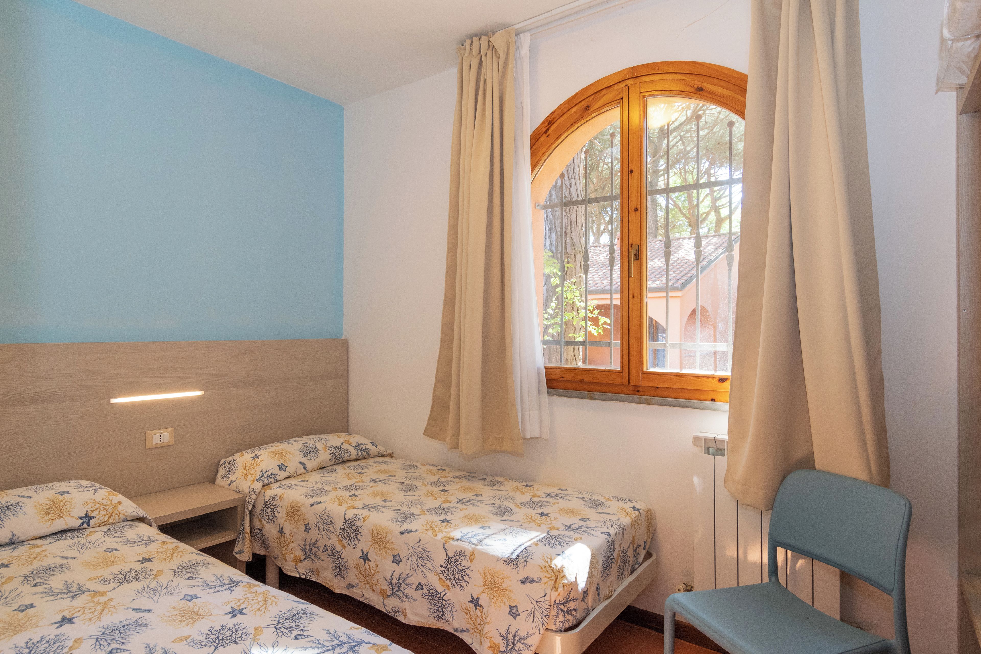 A bedroom with two single beds, a window with curtains, a chair, and a wooden headboard.