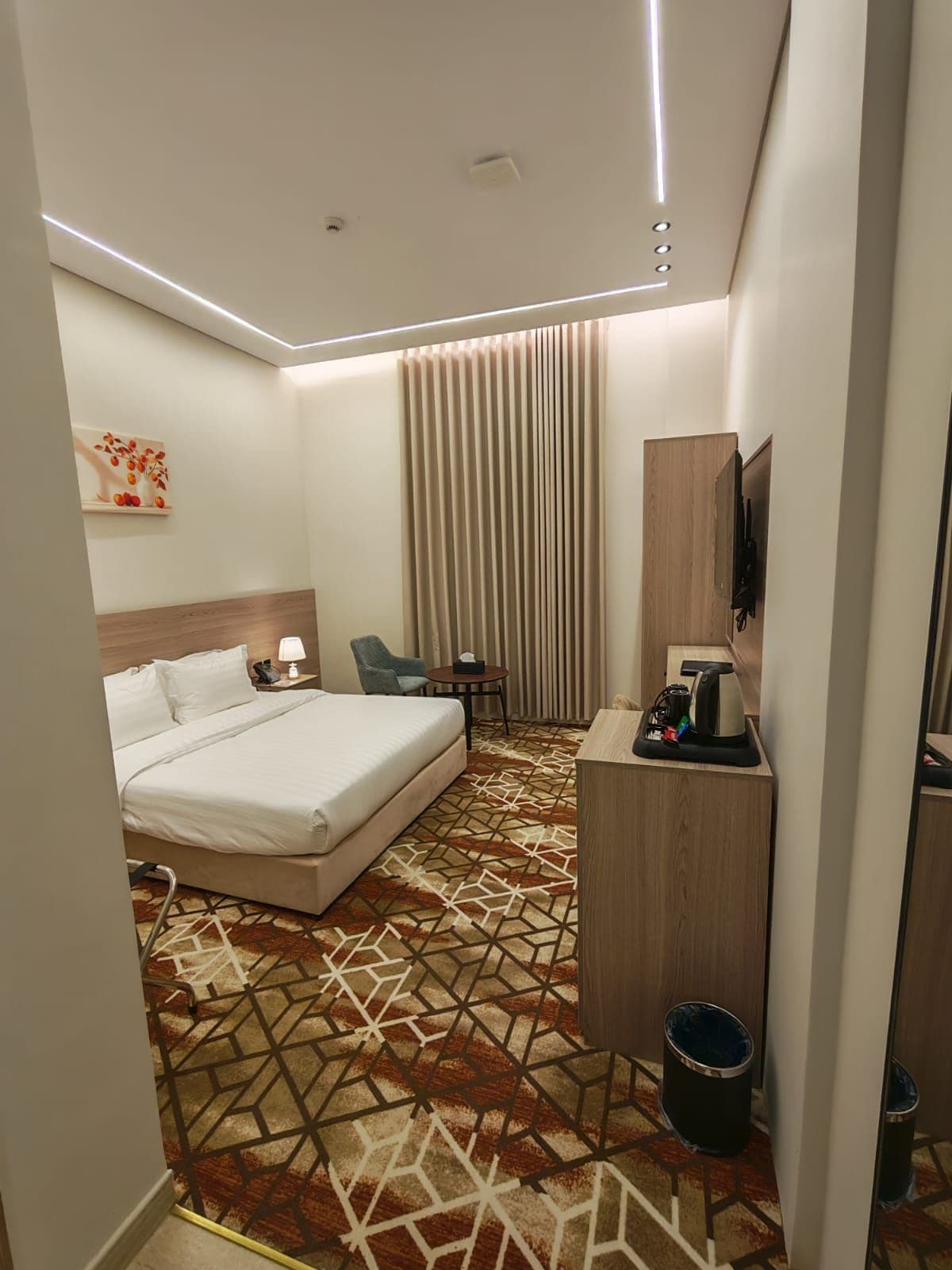 Una habitación de hotel con cama, mesita de noche, lámpara, silla, escritorio y televisor.