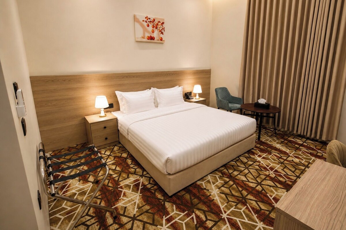 A hotel room with a large bed, bedside tables, a desk, a chair, and a patterned carpet.