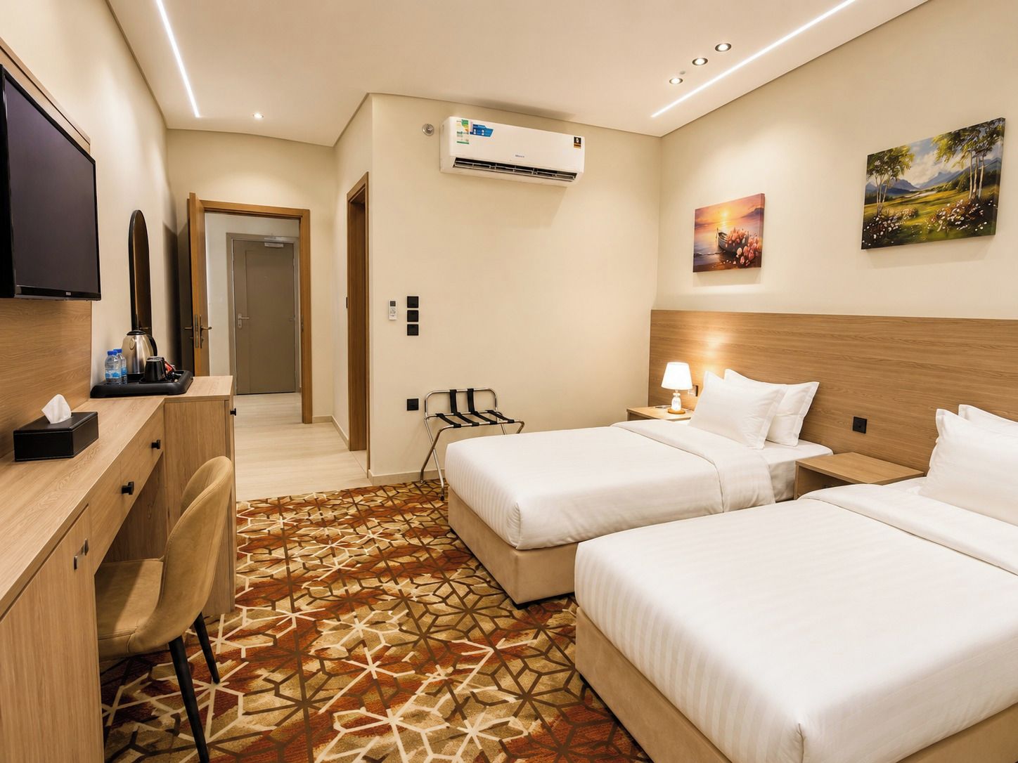 Habitación de hotel con dos camas, un escritorio, una silla, un televisor, aire acondicionado y un cuadro en la pared.
