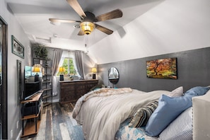 Un dormitorio con cama, escritorio, cómoda, ventilador de techo y ventana con cortinas.