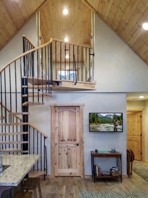 A spacious interior with a wooden staircase leading to a loft area, a dining table, a television mounted on the wall, and a wooden door. - Charming Cabin with Private River Access & Firepit in Ashton, Idaho (Ashton)