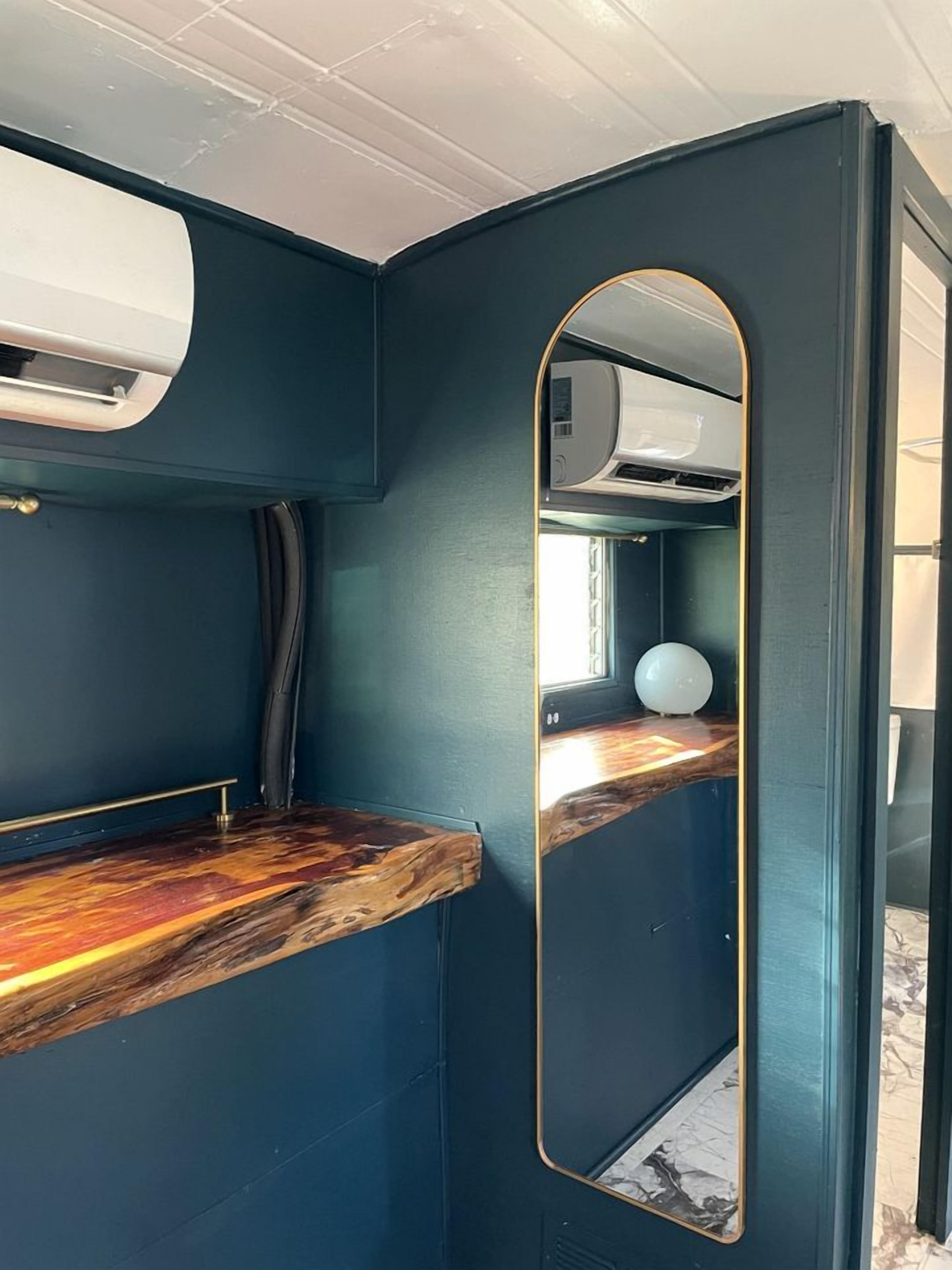 A compact room with a dark blue wall, a wooden countertop, a round mirror, and a ceiling-mounted air conditioning unit.
