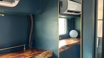 A compact room with a dark blue wall, a wooden countertop, a round mirror, and a ceiling-mounted air conditioning unit.