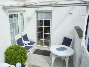 Eine Terrasse mit zwei weißen Stühlen, einem Tisch, einem kleinen grünen Busch und einer Glastür zu einem Innenbereich.