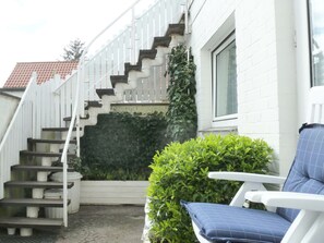 Eine Treppe führt zu einer weißen Tür, ein kleiner Garten mit einer Bank und ein Fenster mit Blick auf ein rotes Ziegeldach.