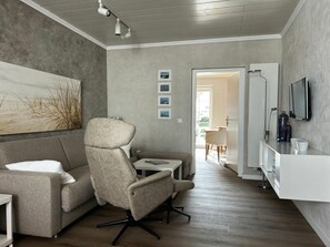 Ein modernes Wohnzimmer mit einem grauen Sofa, einem beigen Sessel, einem kleinen Tisch und einem an der Wand montierten Fernseher.