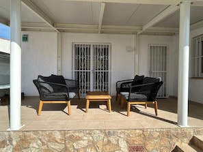 Un espace extérieur aménagé avec une zone salon couverte, comprenant deux chaises en osier noir et une table en bois, des murs blancs et un mur en pierre en bas.