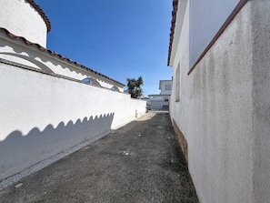 Une ruelle étroite aux murs blancs et au toit recouvert de tuiles, qui mène à une entrée fermée par un portail.