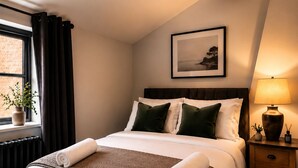 Cama bem arrumada com toalha branca enrolada, manta marrom texturizada e travesseiros verdes. Há um quadro na parede acima da cama, uma janela com cortinas e um abajur em uma mesa de cabeceira de madeira.